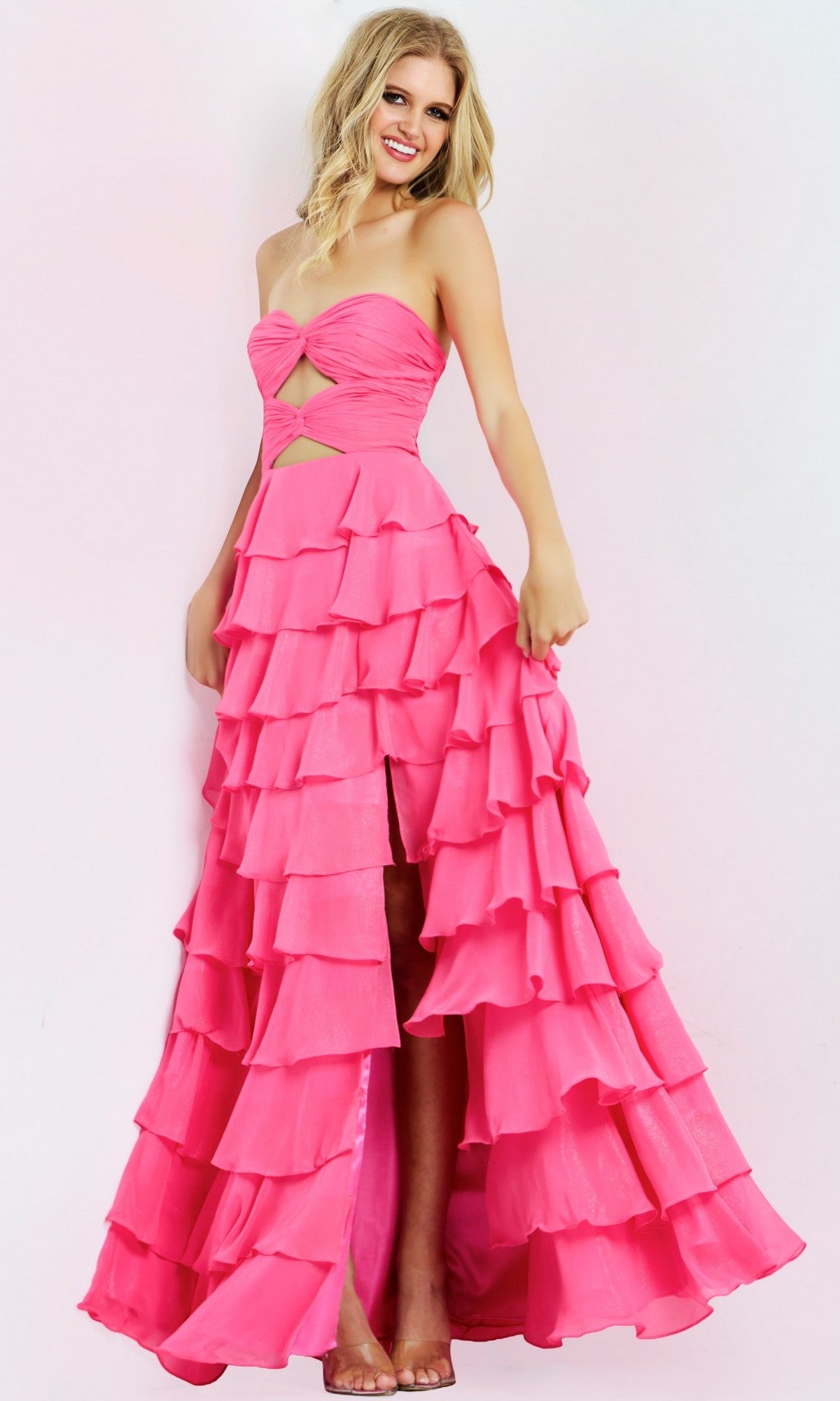 A woman poses against a white background wearing the Jovani 47898 long prom dress, a bright pink strapless gown with a sweetheart neckline, front slit, and multiple chiffon ruffle tiers cascading down the A-line skirt.